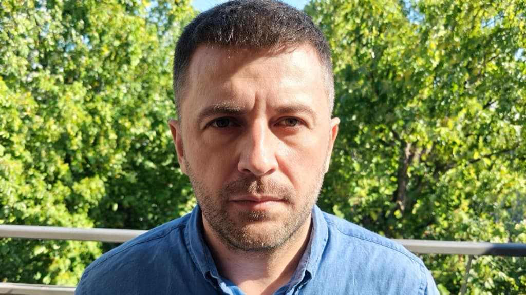 Мужчина 40 лет в синей рубашке на балконе летом, яркое солнце сверху, резкие тени на лице, позади зелень деревьев